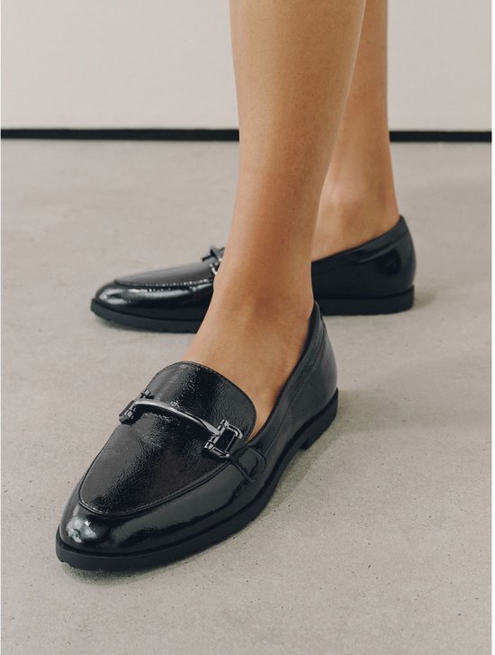 Een vrouw draagt zwarte lakleren loafers met zilverkleurige gespen.