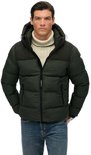 Man met beige coltrui en donkergroene, gewatteerde jas met capuchon, staand met handen in zakken.