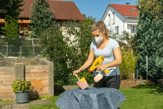 Een vrouw met mondkapje gebruikt een gele verfspuit op een grijze steen in de tuin.