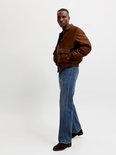 Man met bruine jas en wijde blauwe jeans, staand.