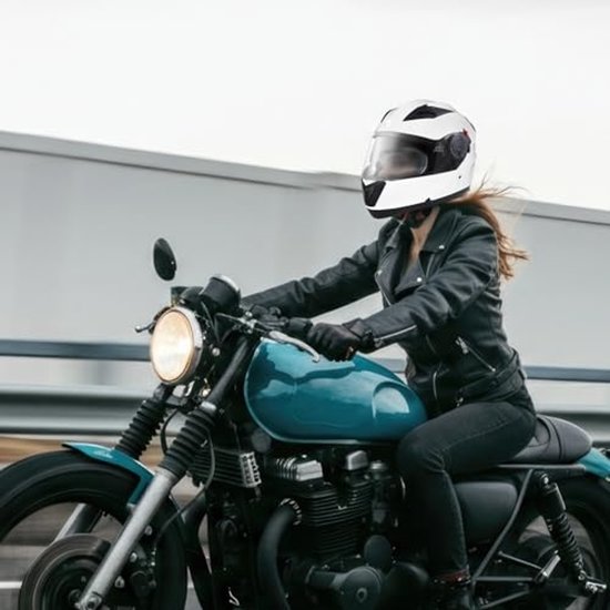 Vrouw op een klassieke motorfiets draagt een witte systeemhelm, zwart leren jack en spijkerbroek.