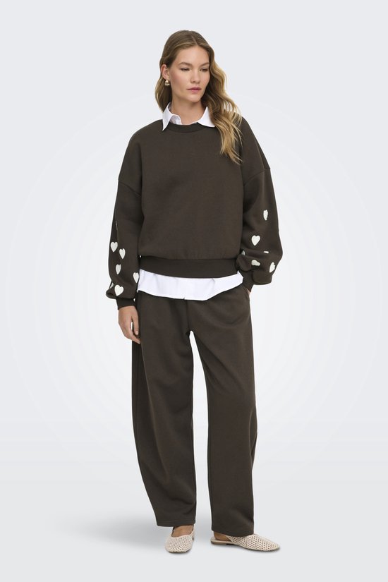 Donkerbruine sweater met witte hartjes op de mouwen, gedragen door een model met een witte blouse eronder.