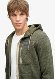 Close-up van een olijfgroene fleece hoodie met beige rits en Superdry & Co-badge.