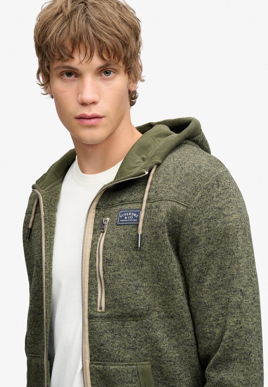 Close-up van een olijfgroene fleece hoodie met beige rits en Superdry & Co-badge.