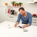 Man lacht terwijl hij een IQ Fit spel speelt in een keuken. Hij houdt een stuk van de puzzel vast en het spel ligt op tafel.