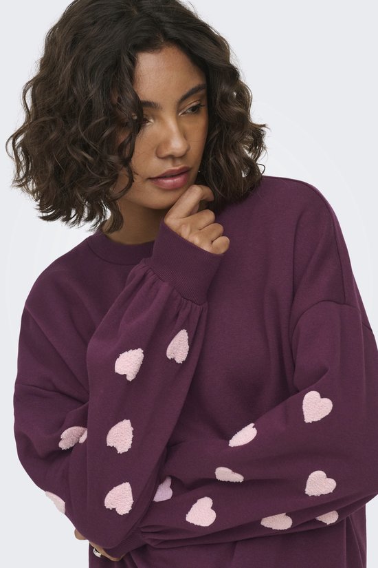 Donkerpaarse sweater met geborduurde lichtroze hartjes op de mouwen. Model draagt de sweater.