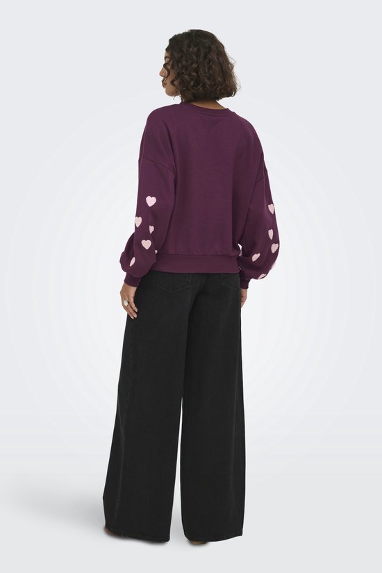 Zijaanzicht van een donkerpaarse sweater met lichtroze hartjes op de mouwen, gedragen door een model met wijde zwarte broek.