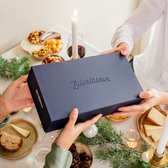 Twee personen geven elkaar een blauwe kerstcadeaubox van Zuivelhoeve aan een gedekte tafel met een kerstborrelplank.