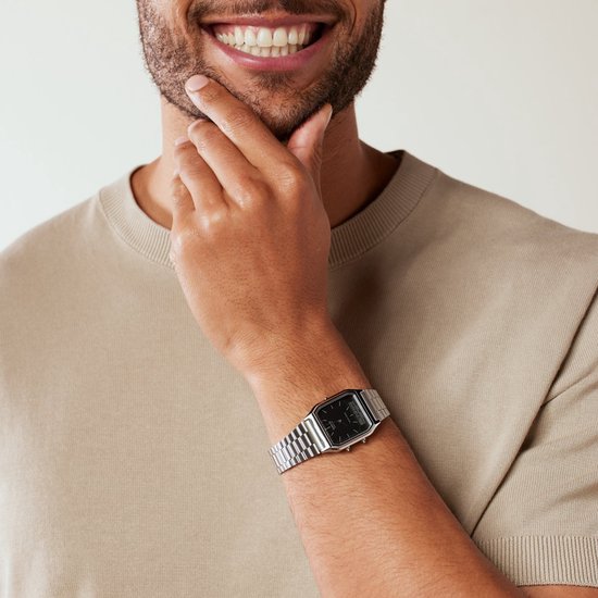 Zilverkleurig metalen horloge met zwart vierkant display om de pols van een man met een beige trui.