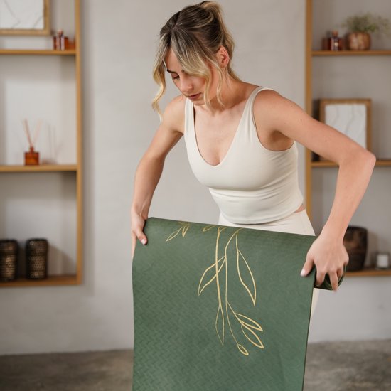 Vrouw in beige outfit rolt een donkergroene yogamat met gouden plantenprint op.