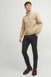 Man in een beige bomberjack en zwarte chino broek, staand.