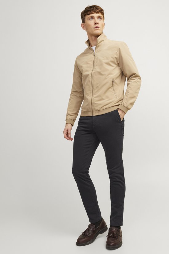 Man in een beige bomberjack en zwarte chino broek, staand.
