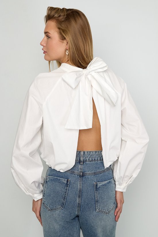 Model draagt witte blouse met open rug en strik, met lange mouwen en hoge hals, gecombineerd met spijkerbroek.