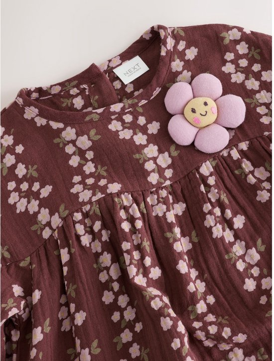 Bruine jurk met lange mouwen en bloemenprint. Op de borst zit een applicatie van een lachende bloem.