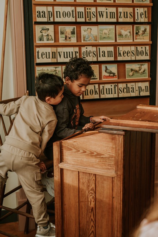 Twee jongens staan bij een houten bureau met schoolplaten met letters en afbeeldingen van dieren en mensen.