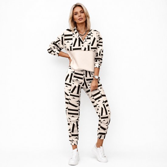 Vrouw staat op witte achtergrond in crème-kleurig trainingspak met zwart logo-print, beige bovenstuk.