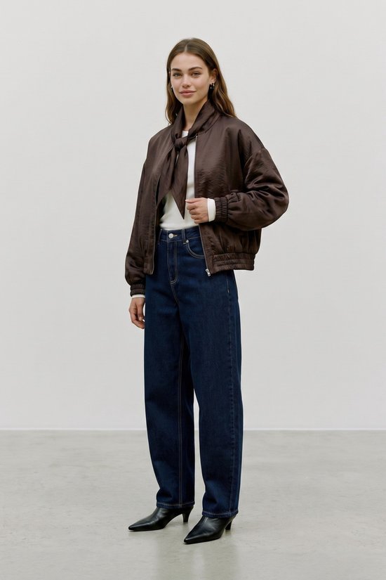 Model in een bruin satijnen bomberjack met strik detail, witte top en jeans.