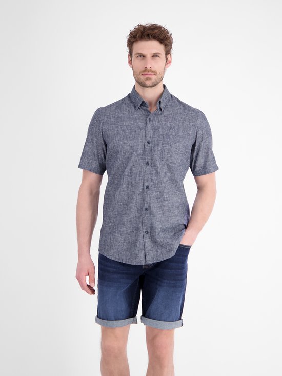 Man met donkerblauw kortademig overhemd en denim short, vooraanzicht met hand in zak.