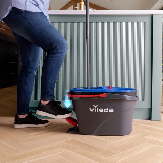 Persoon met spijkerbroek en sneakers gebruikt een Vileda spinmop met pedaalemmer in een keuken.