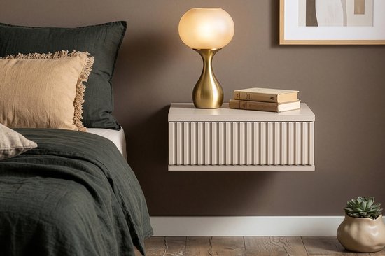 Beige hangend nachtkastje met verticale ribbels op ladefront, naast een bed met kussens, lamp en plant.