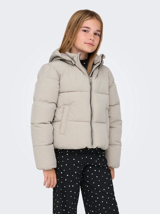 Meisje met lange blonde haren draagt een beige korte pufferjas en een zwarte broek met witte stippen.