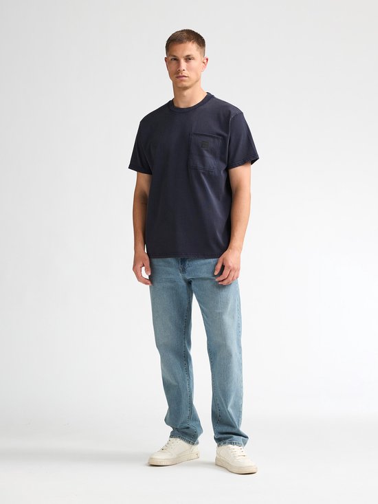 Model draagt een donkerblauw T-shirt met borstzak, jeans en sneakers.