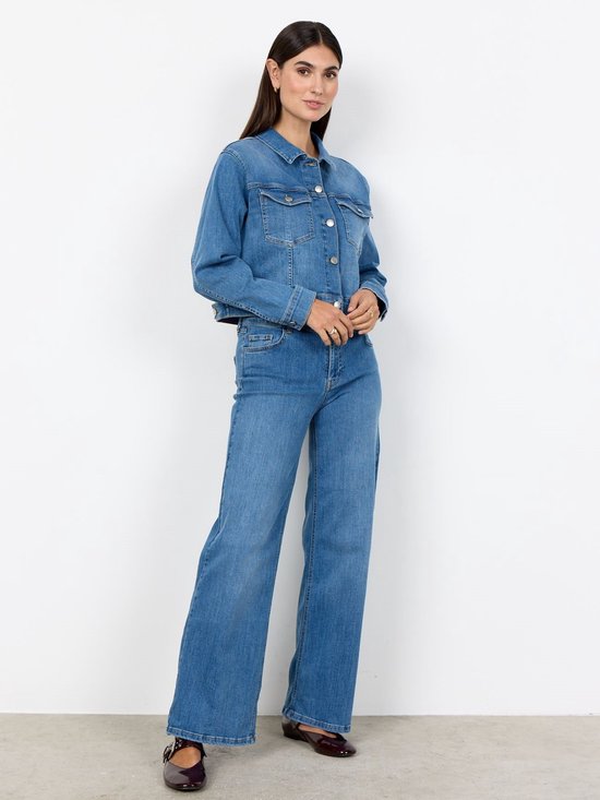 Model draagt een lichtblauwe spijkerjas en bijpassende jeans met wijde pijpen en donkerrode platte schoenen.