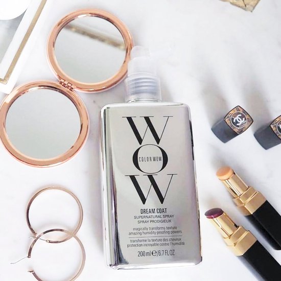 Zilverkleurige fles "DREAM COAT SUPERNATURAL SPRAY" op een marmeren oppervlak, met make-up en spiegels.