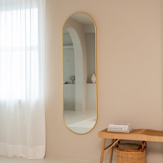 Gouden ovale spiegel aan een beige muur, naast een houten bankje met boeken en een mand.