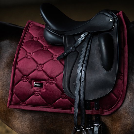 Donkerrode, gepofte dressuur deken met diamantpatroon onder een zwart zadel op een paard.