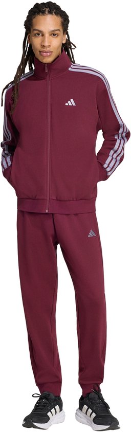 Model in een bordeauxrode trainingsbroek en trainingsjack met lichtpaarse strepen op de mouwen. Adidas logo op de borst en broek.