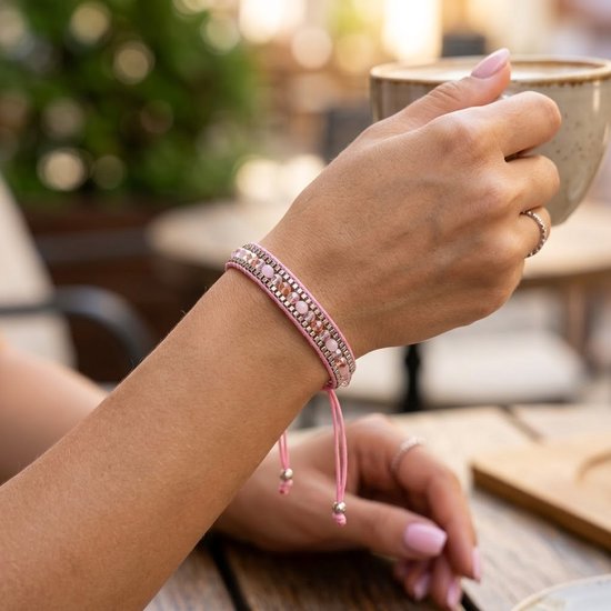 Damesarm met roze geweven armband met facet geslepen glaskralen, aan een verstelbaar koord. Houdt een kop koffie vast.