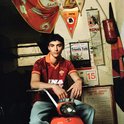Jonge man in rood AS Roma voetbalshirt met Ina logo, lichtblauwe broek, zittend op oranje scooter.