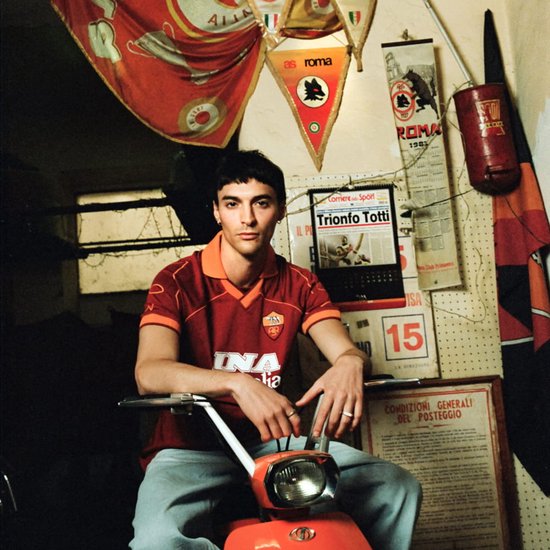 Jonge man in rood AS Roma voetbalshirt met Ina logo, lichtblauwe broek, zittend op oranje scooter.