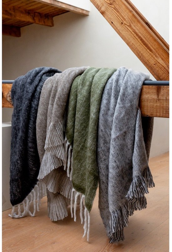 Drie gebreide plaids met franjes, in donkergrijs, beige en groen, hangend over een houten balk.