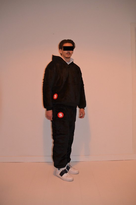 Een man staat frontaal, met geblurde ogen, gekleed in een oversized zwart trainingspak met capuchon en een rood S-logo op de borst en linkerbroekspijp. De capuchon is omgeslagen, met witte voering zichtbaar. Hij draagt witte sneakers.