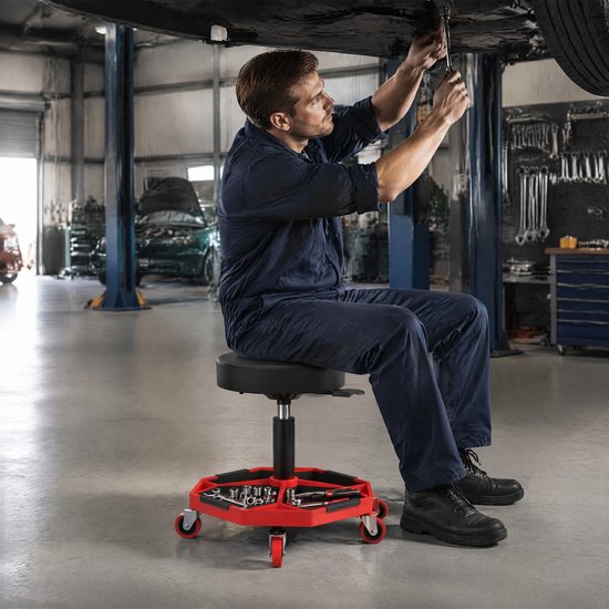 Man in blauw overall zittend op een werkplaatskruk met gereedschapsbak onder een auto.