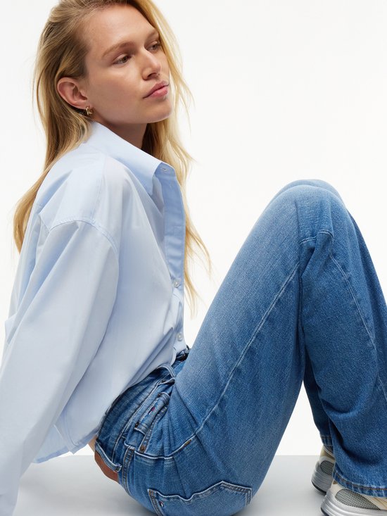 Model zit, draagt een rechte spijkerbroek in middelblauwe wassing en een lichtblauwe blouse.