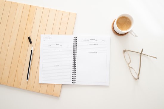 Open notitieboek, kopje koffie, bril, houten ondergrond en potlood op een witte tafel.