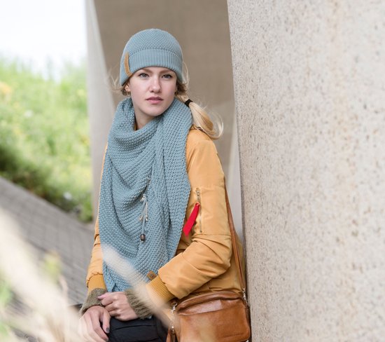 Model met lichtblauwe gebreide sjaal en bijpassende muts, draagt een okergele jas.