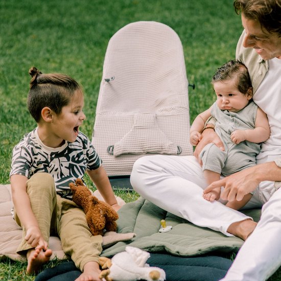 Man zit in het gras met twee kinderen en een beige wipstoel op de achtergrond.