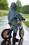 Kind in een lichtgroen regenpak met visgraatpatroon, zittend op een loopfiets in de regen.