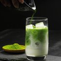 Matcha wordt in een glas met melk en ijs gegoten, met een lepel matcha poeder ernaast.