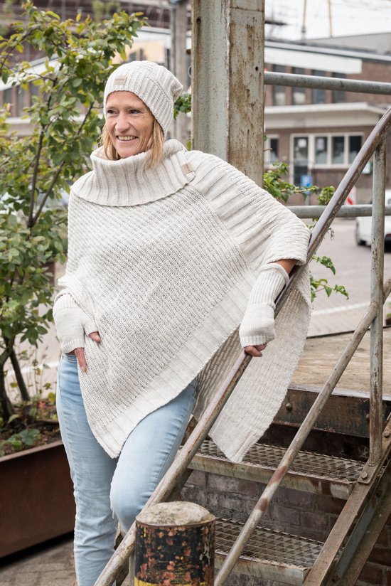 Lichtbeige gebreide poncho met hoge sjaalkraag en bijpassende muts, gedragen door een vrouw met blond haar op een trap.