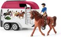 Bruine paard met ruiter in blauwe jas, naast witte trailer met bordeauxrode bovenkant en paardenafbeeldingen.