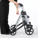 Vrouw in zwarte broek duwt een opgevouwen grijze rollator met handvatten en remhendels.