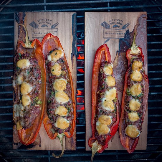 Twee cederhouten planken met rode paprika's gevuld met vlees en kaas, op een barbecue.