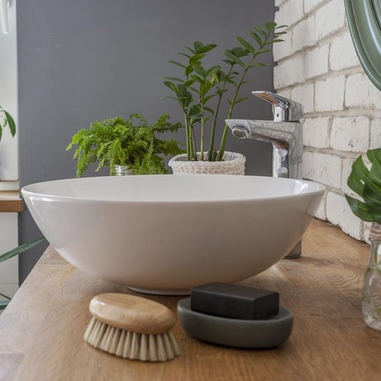 Lavabo rond en céramique blanche sur un comptoir en bois, avec un robinet, des plantes et un morceau de savon.