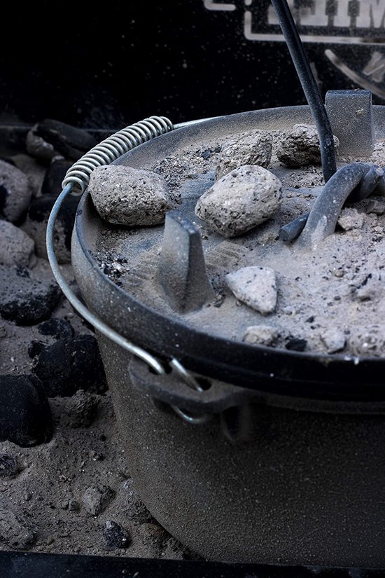 Gietijzeren Dutch oven met deksel bedekt met as en kolen, op een rooster.