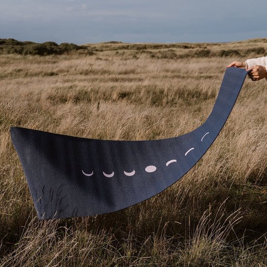 Donkerblauwe yogamat met een print van maanfasen, gehouden in een weiland met hoog gras.
